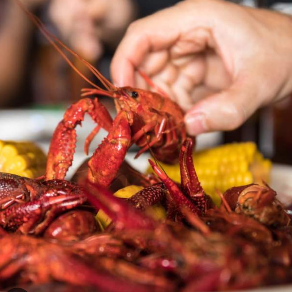 A hand reaching for a bright red crawfish from a pile of seafood and corn on the cob.